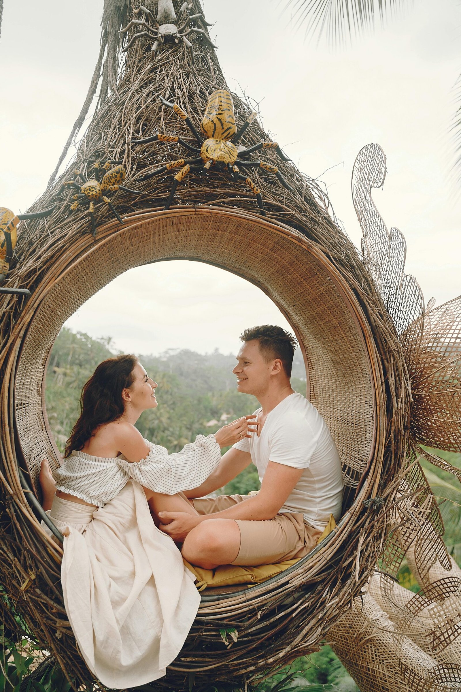 Side view full body barefoot content young couple in white casual clothes sitting in wicker hanging swing and looking at each other with love while spending honeymoon in tropical resort